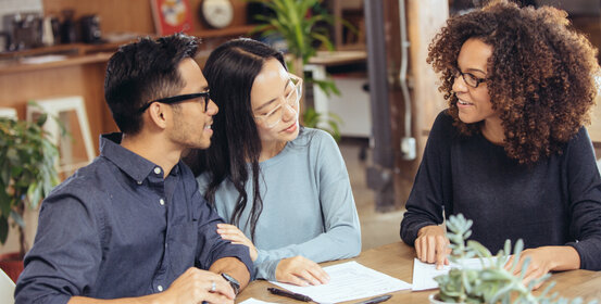 A couple reviews documents with a consultant. They sit at a table in a casual office setting, engaged in discussion.