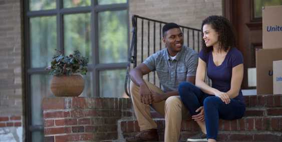 A man and woman sitting on their front porch with moving boxes around them