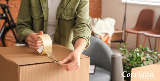 A person tapes a cardboard box shut using clear packing tape. The action takes place in a home, amidst moving boxes and furniture. Corrigan Housing Solutions.