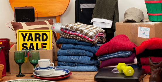 Clothing and household goods are being displayed for sale at a yard sale. A sign reads "YARD SALE". A box is labeled "Books 50¢ each".