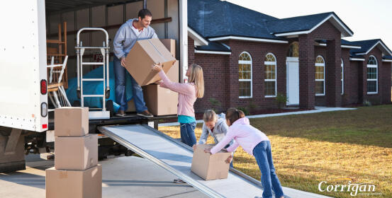 A family unloads cardboard boxes from a moving truck, using a ramp, into the yard of a brick house. Corrigan.