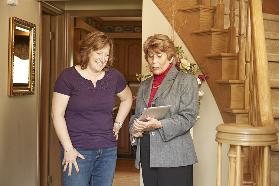 Two women review a tablet in a home's entryway. One woman wears a purple shirt, the other a gray blazer. A wooden staircase is visible.