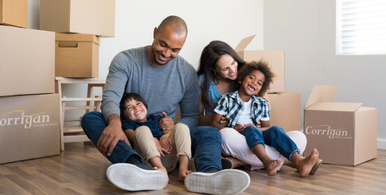 A family sits on a hardwood floor amidst moving boxes. The parents playfully hold their two young children. The boxes are labeled "Corrigan Moving Systems."